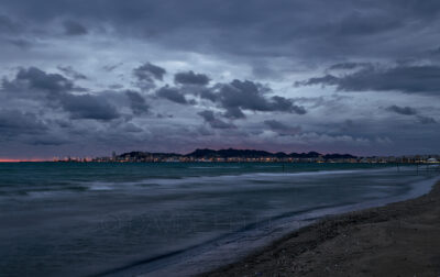This landscape photograph captures a moody scene of storm clouds hovering over the sunset of Durres city in Albania. The photograph features the striking contrast between the dark clouds and the choppy sea, with the nighttime lights of the Durres port visible in the distance. The overall atmosphere is both ominous and captivating, making for a truly stunning image.