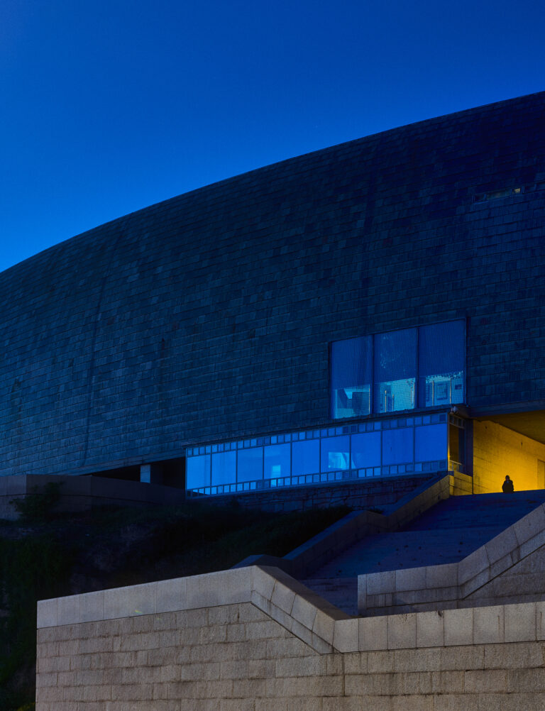 Curved Stone Building With Reflective Glass Windows, Illuminated Entrance, And Lone Figure At Dusk; Architectural Exterior Photo.